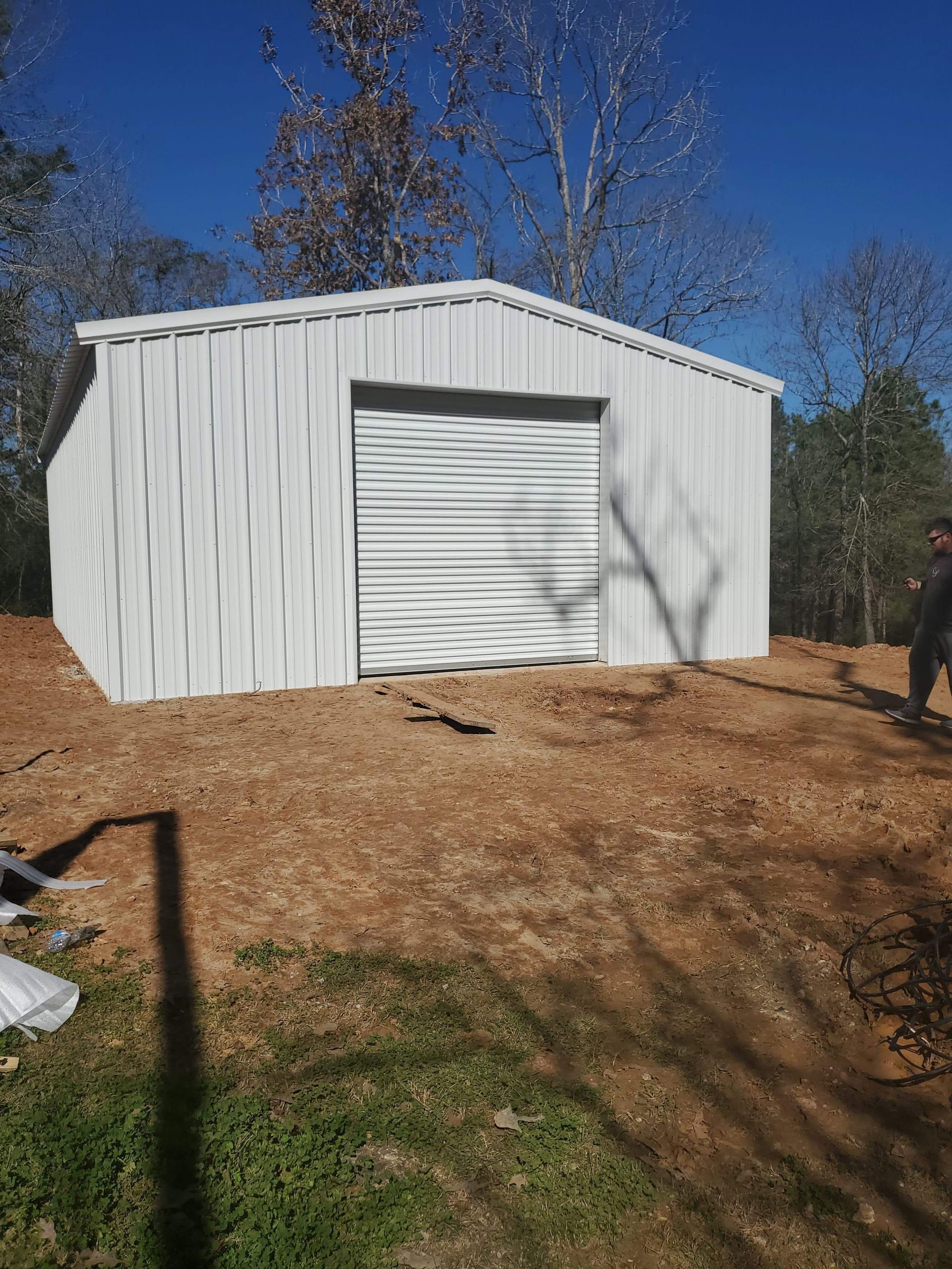White metal shed with a closed garage door, on a dirt patch, with a blue sky background.