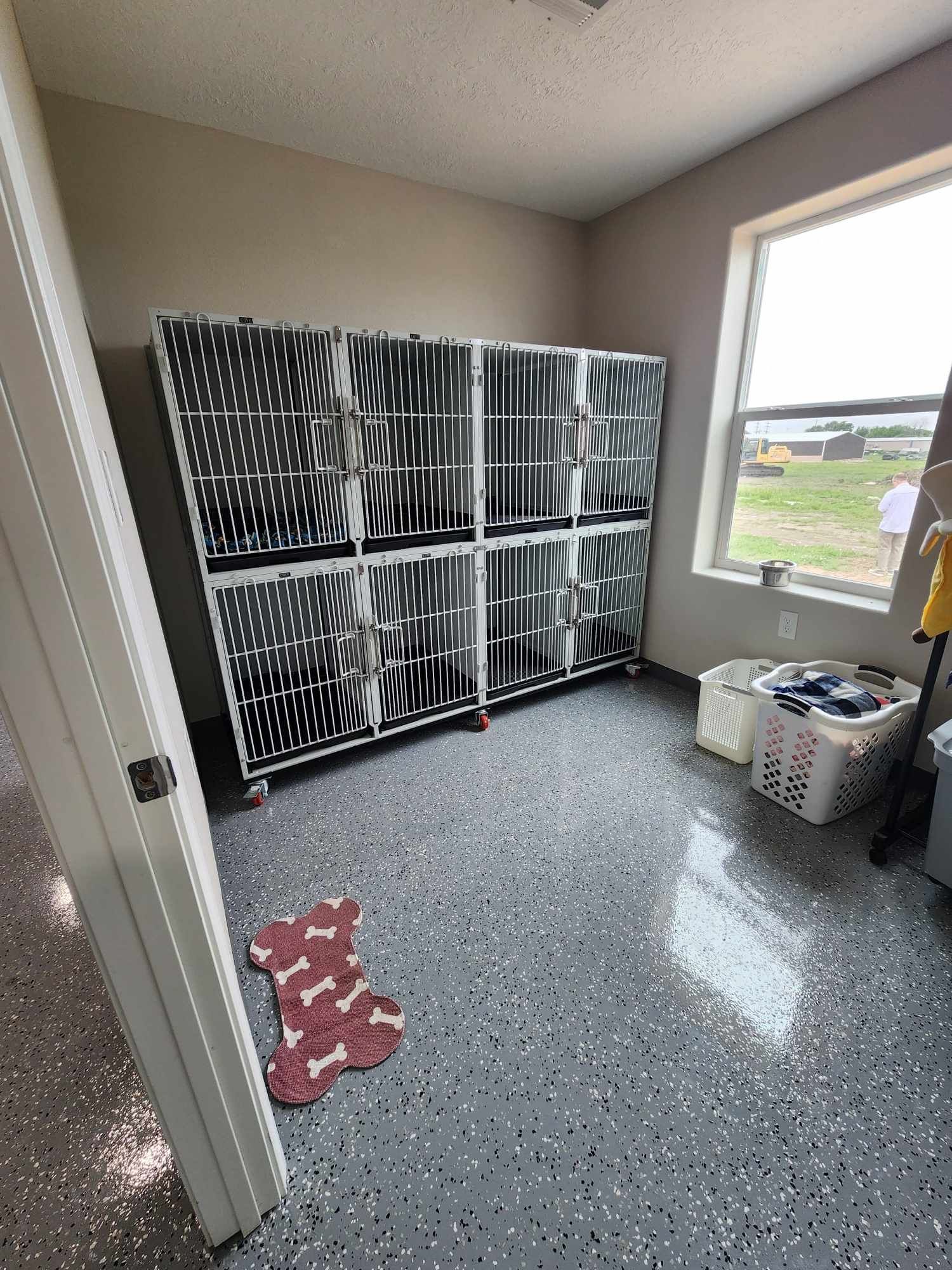 A room with dog kennels, a window, and a speckled floor. A door and dog bed are visible.