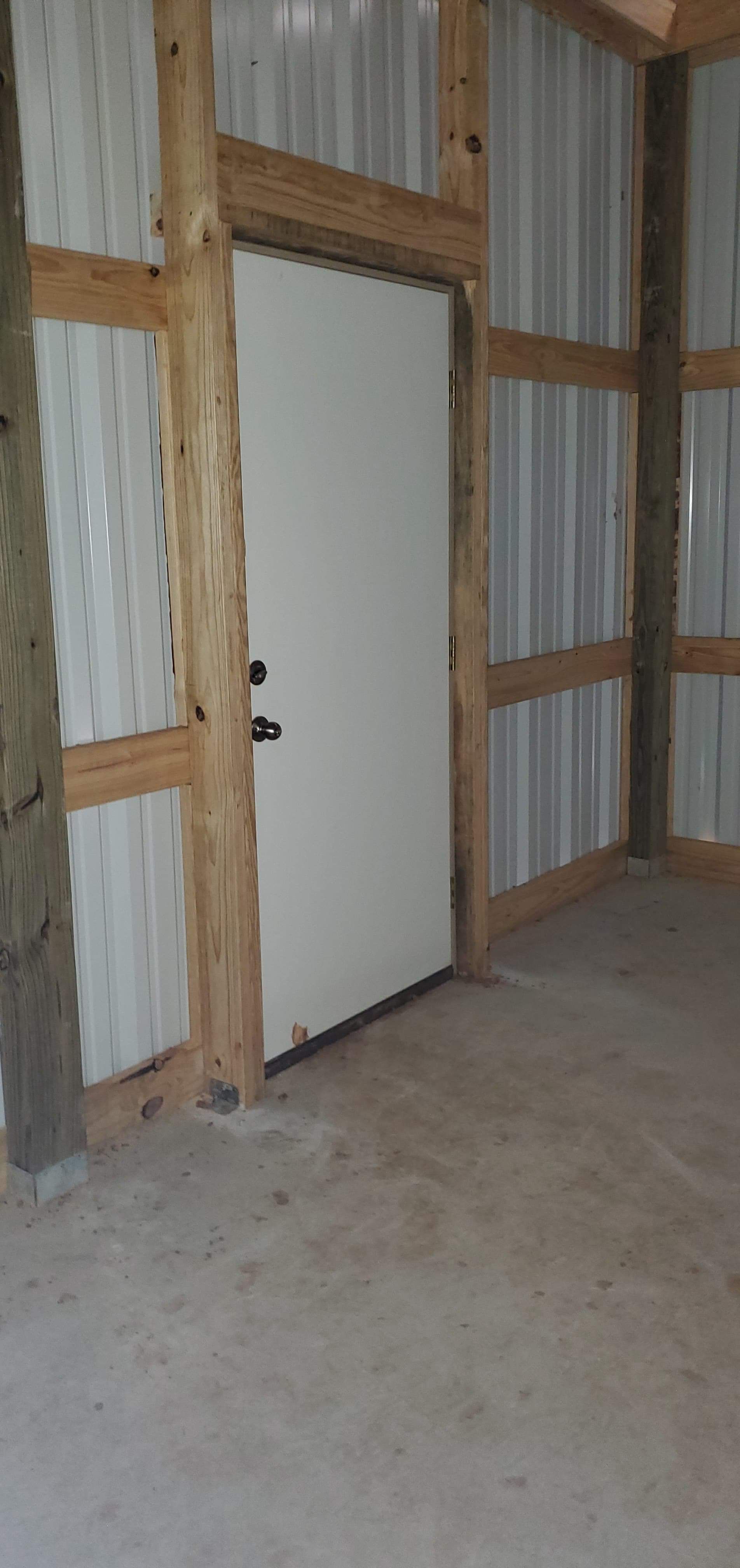 Interior view of a barn with a closed white door framed by wooden beams and metal siding.