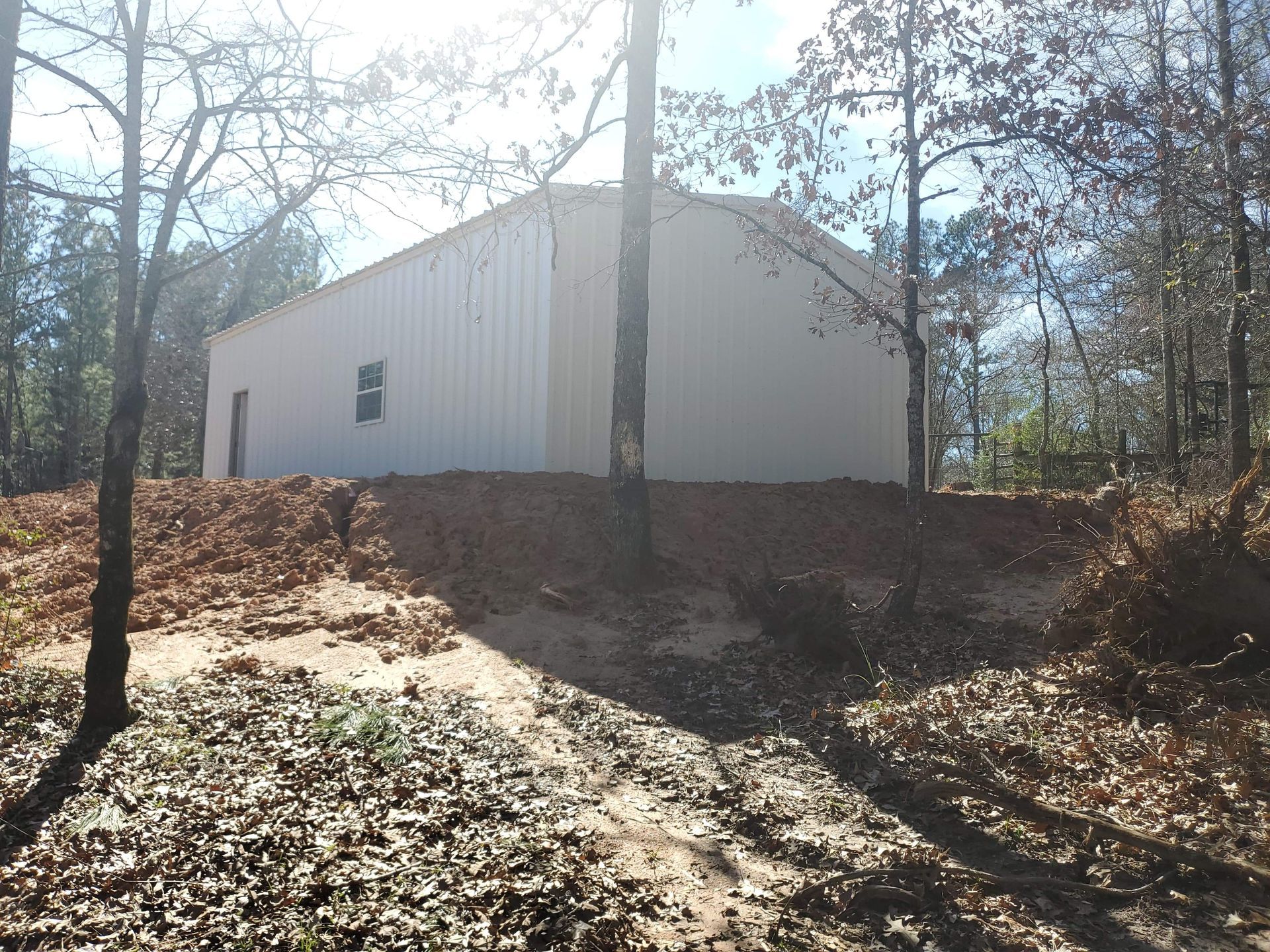 White metal building on a dirt mound in a wooded area; daytime with bare trees and fallen leaves.