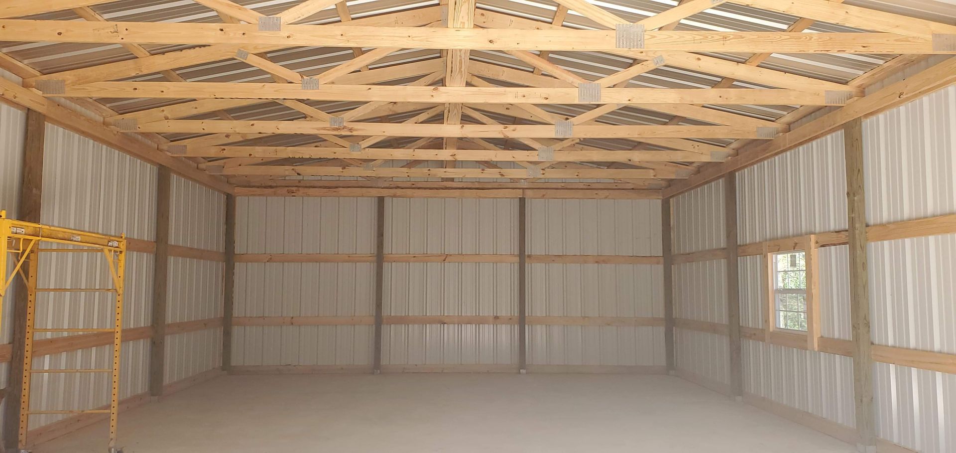 Interior view of an unfinished building with exposed wooden beams, metal siding, and a dirt floor.