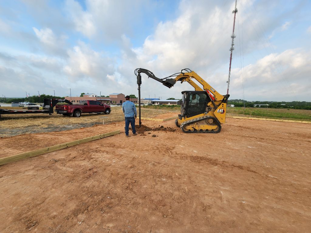 A worker operates a yellow skid steer drilling into the ground, a red truck and building in the background.