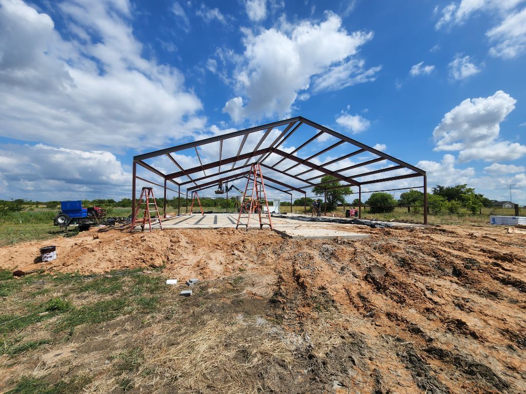 Steel frame of a building under construction against a blue sky. Dirt and grass in foreground.