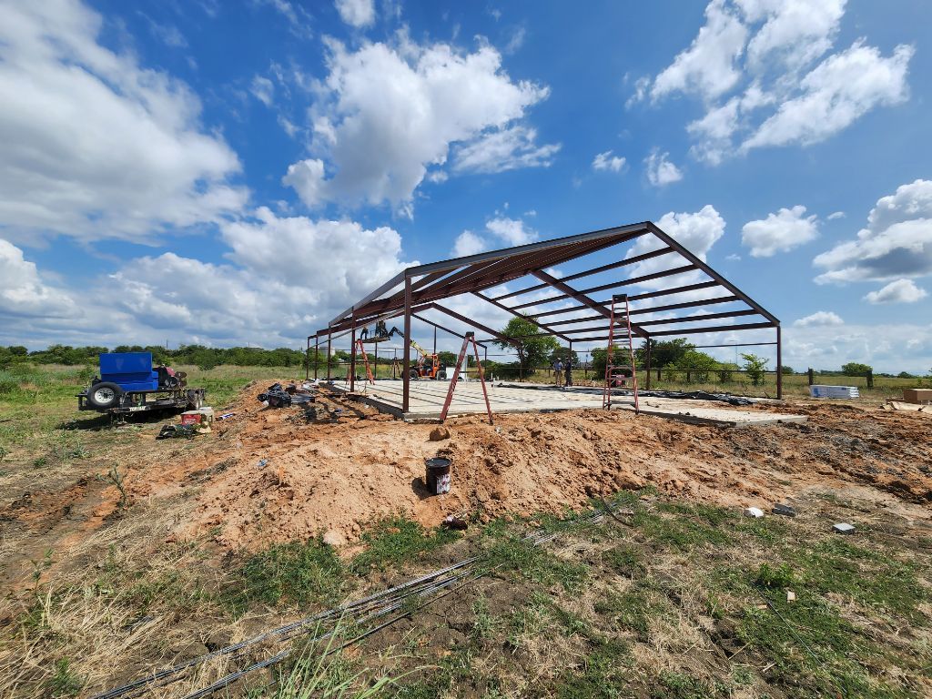 Steel frame of a building under construction on a dirt lot, blue sky with clouds in the background.
