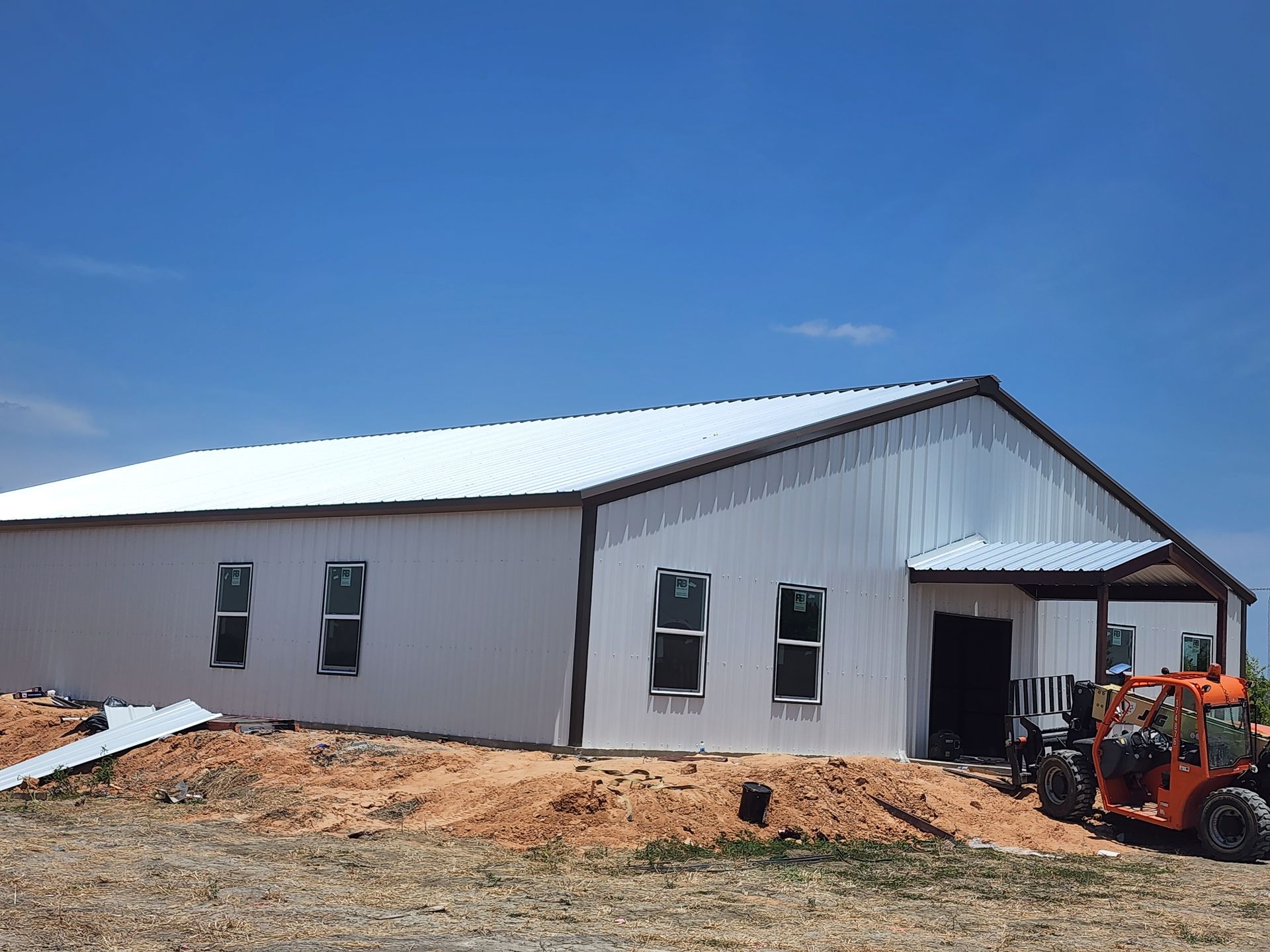 White metal building with brown trim, windows, and a front overhang under a blue sky; construction site.