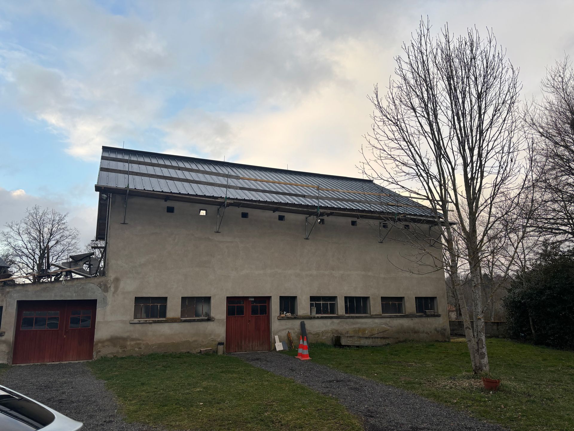Bâtiment gris au toit en pente et à la porte rouge. Ciel couvert et arbres nus.