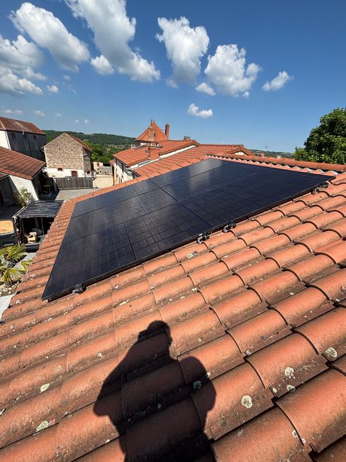 Panneaux solaires installés sur un toit en tuiles de terre cuite, avec l'ombre d'une personne devant. Ciel bleu.