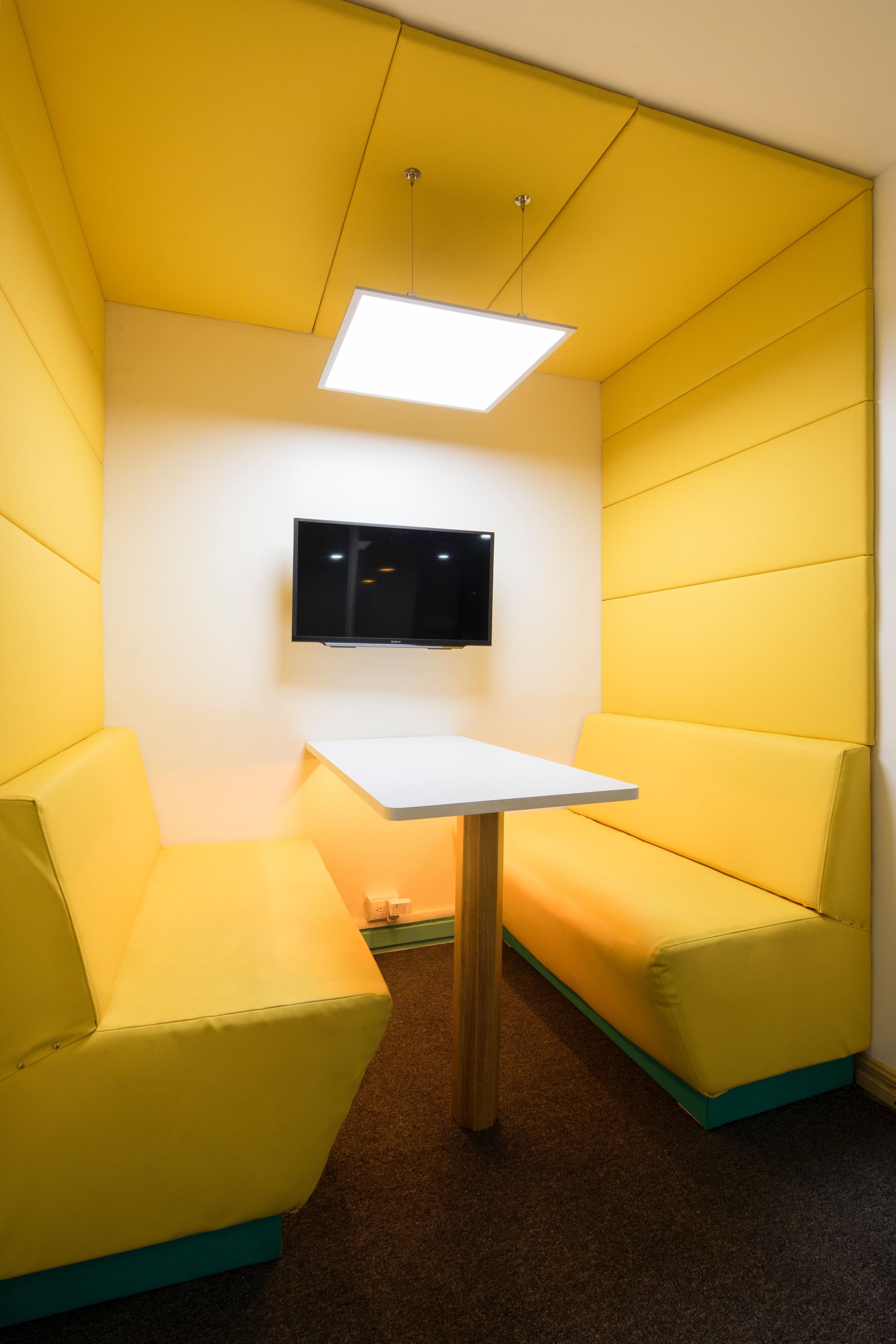 Een gele overlegplek met banken, een tafel en een tv aan de muur