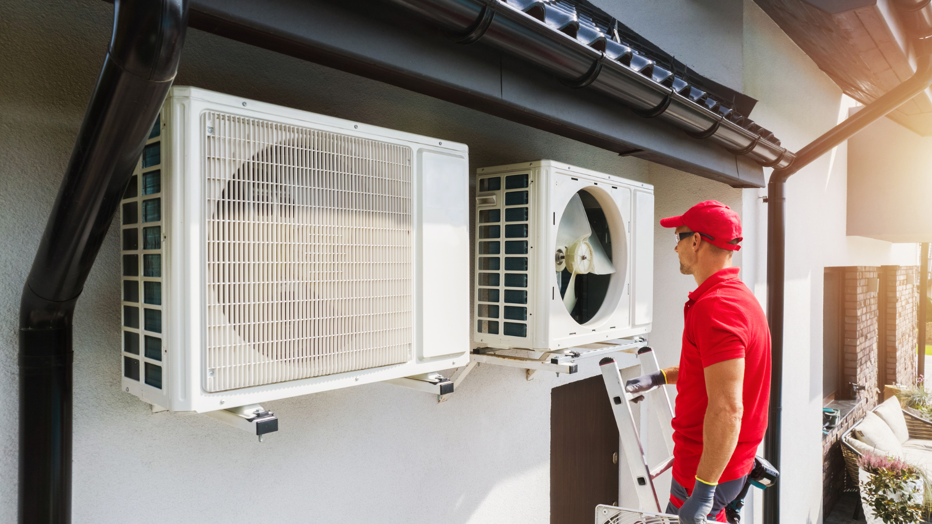 HVAC technician in red shirt and cap inspects AC units mounted on a building's wall.