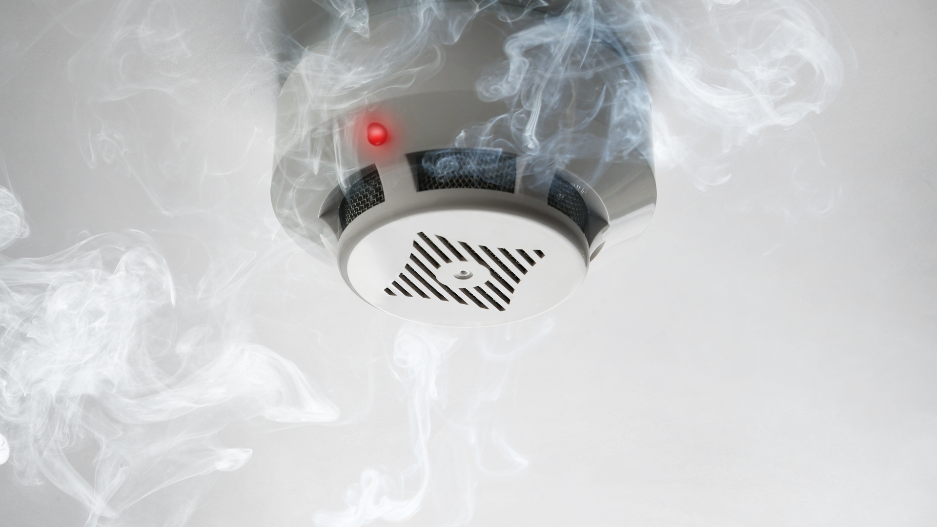 Smoke rising around a ceiling-mounted smoke detector with a red light illuminated.