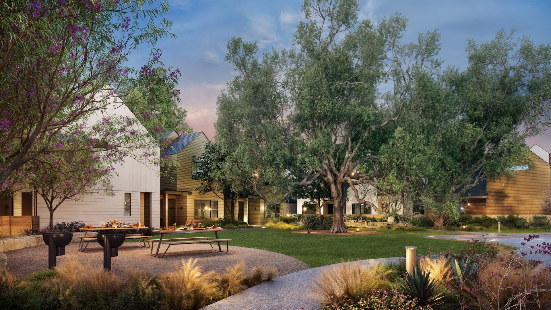 Park with trees and buildings. Picnic tables on gravel path. Green grass, warm lighting.