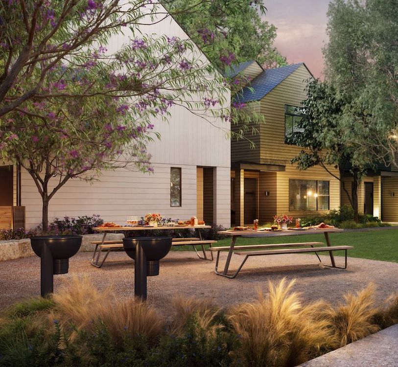 Outdoor dining area with picnic tables, decorative fire pits, and houses in the background, with trees and landscaping.
