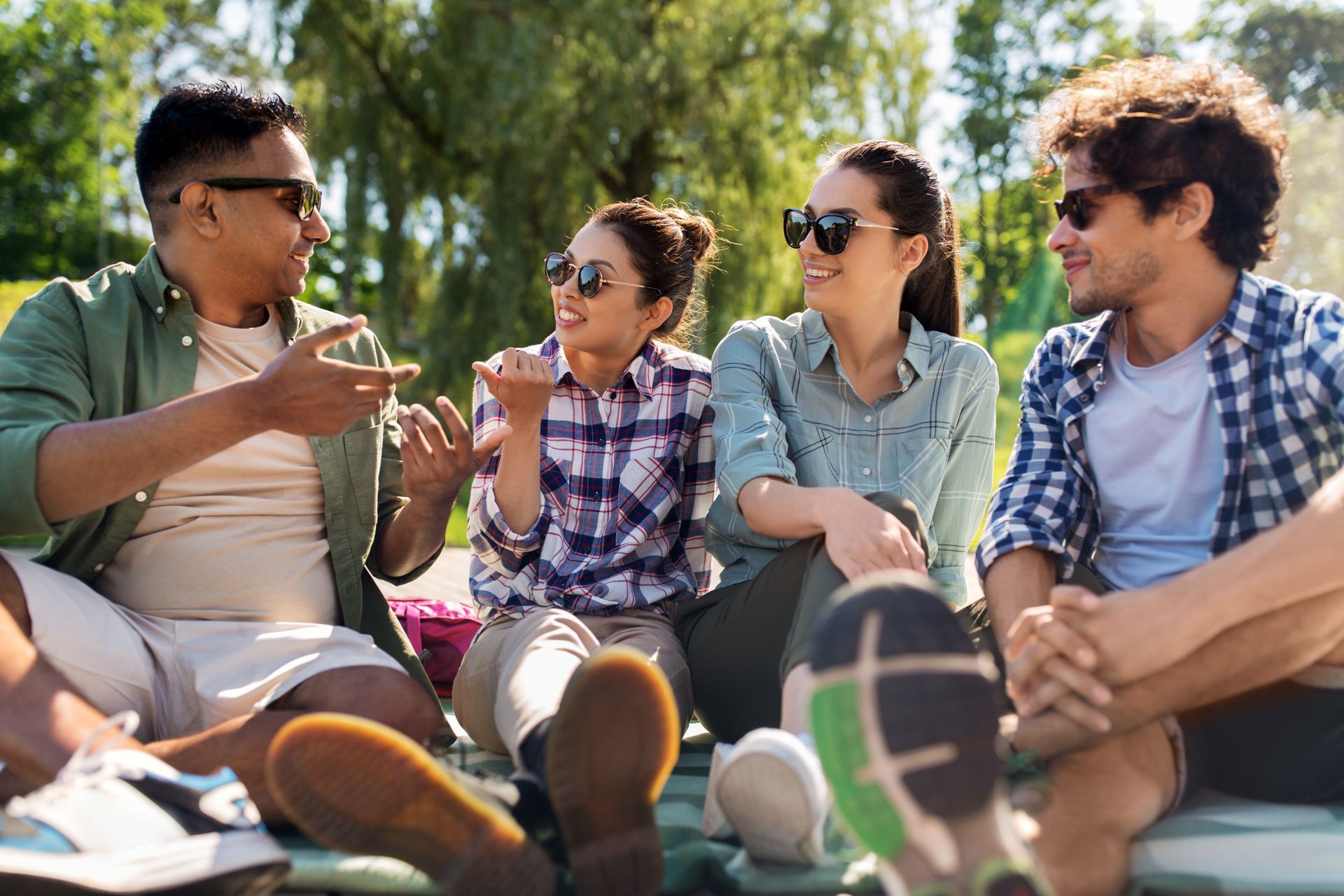 Four friends wearing sunglasses sit and talk outside.