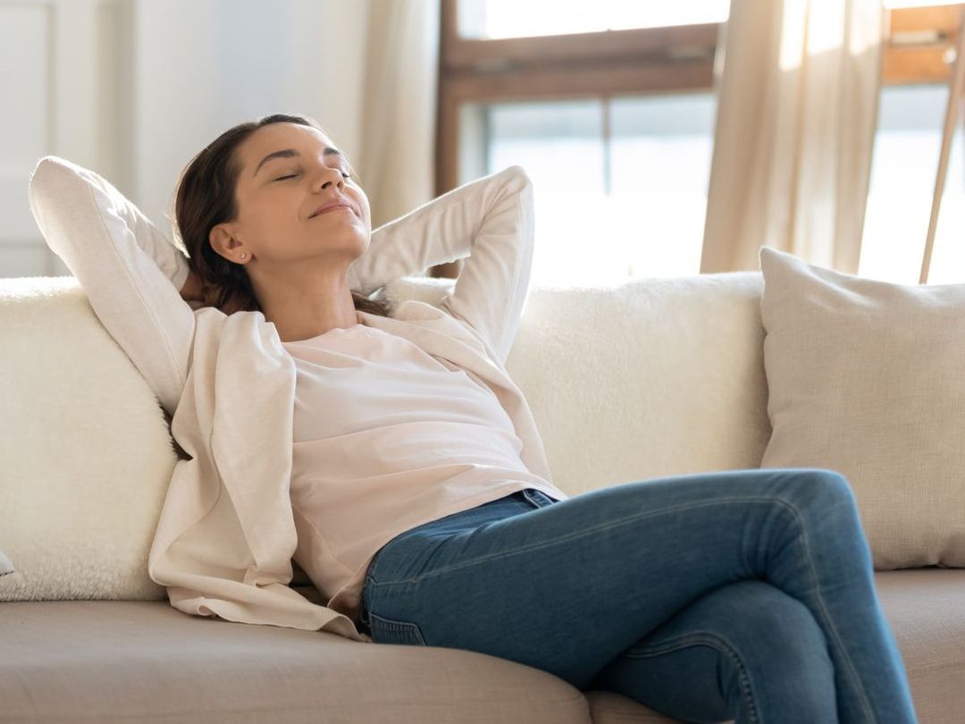 Woman relaxing on a couch, eyes closed, arms behind head, sunlight streaming in.