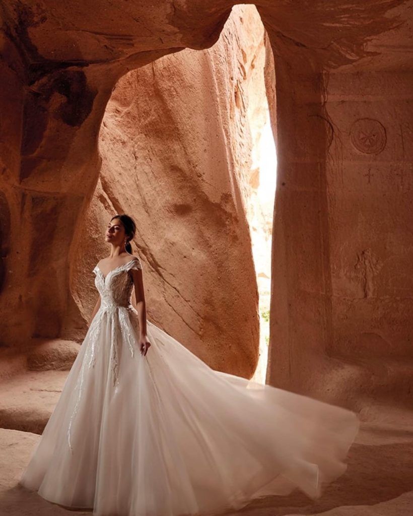 Woman in white wedding dress with train stands in a rock cave, looking upwards.