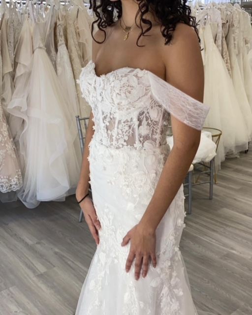 Woman in a white off-the-shoulder wedding dress with floral details and a mermaid silhouette, in a bridal shop.