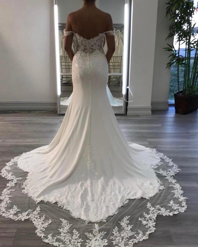 Woman in white off-the-shoulder wedding dress with long train, standing in front of mirror.