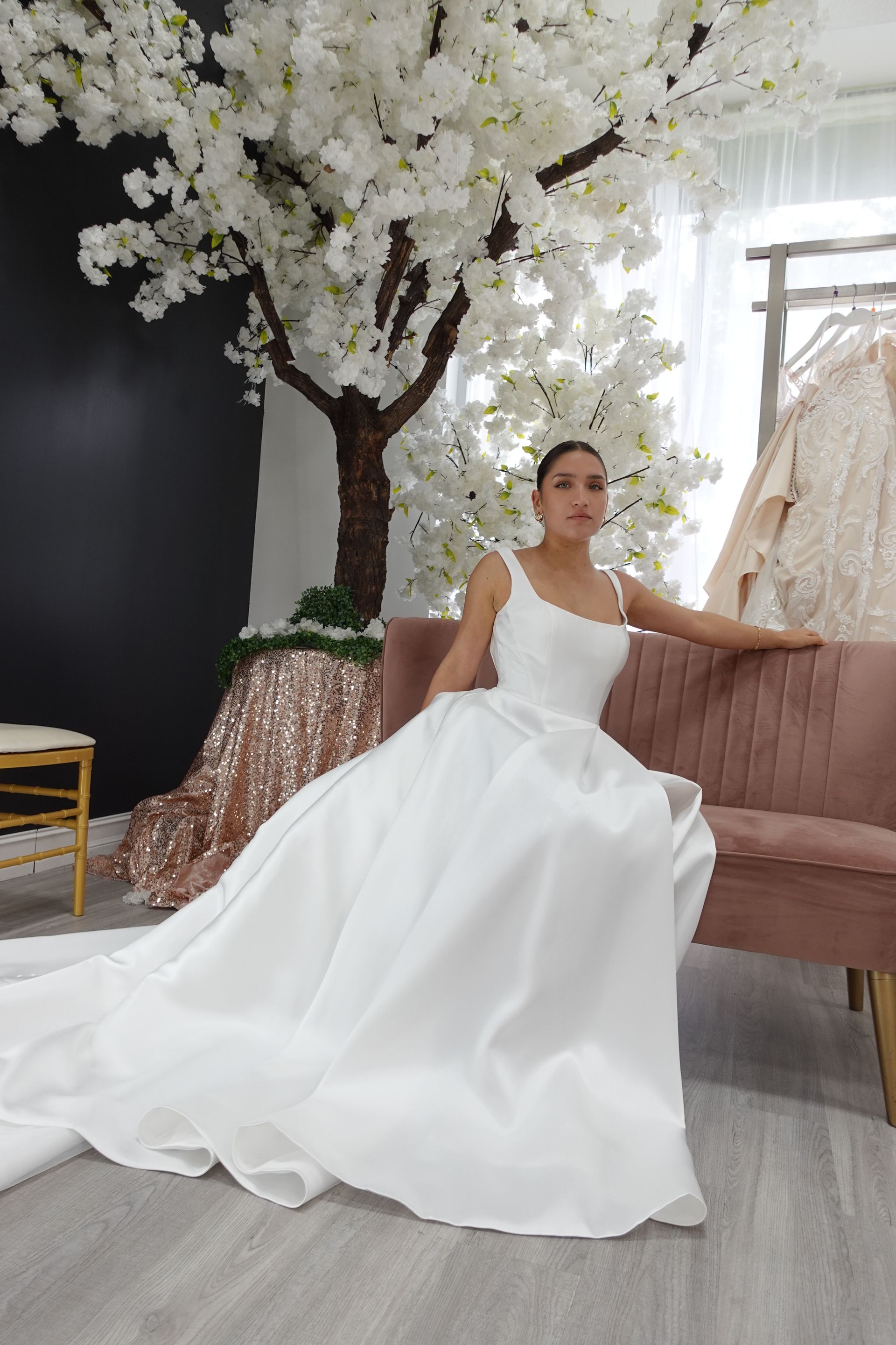 Woman in white wedding dress sits on pink couch under a blooming tree.