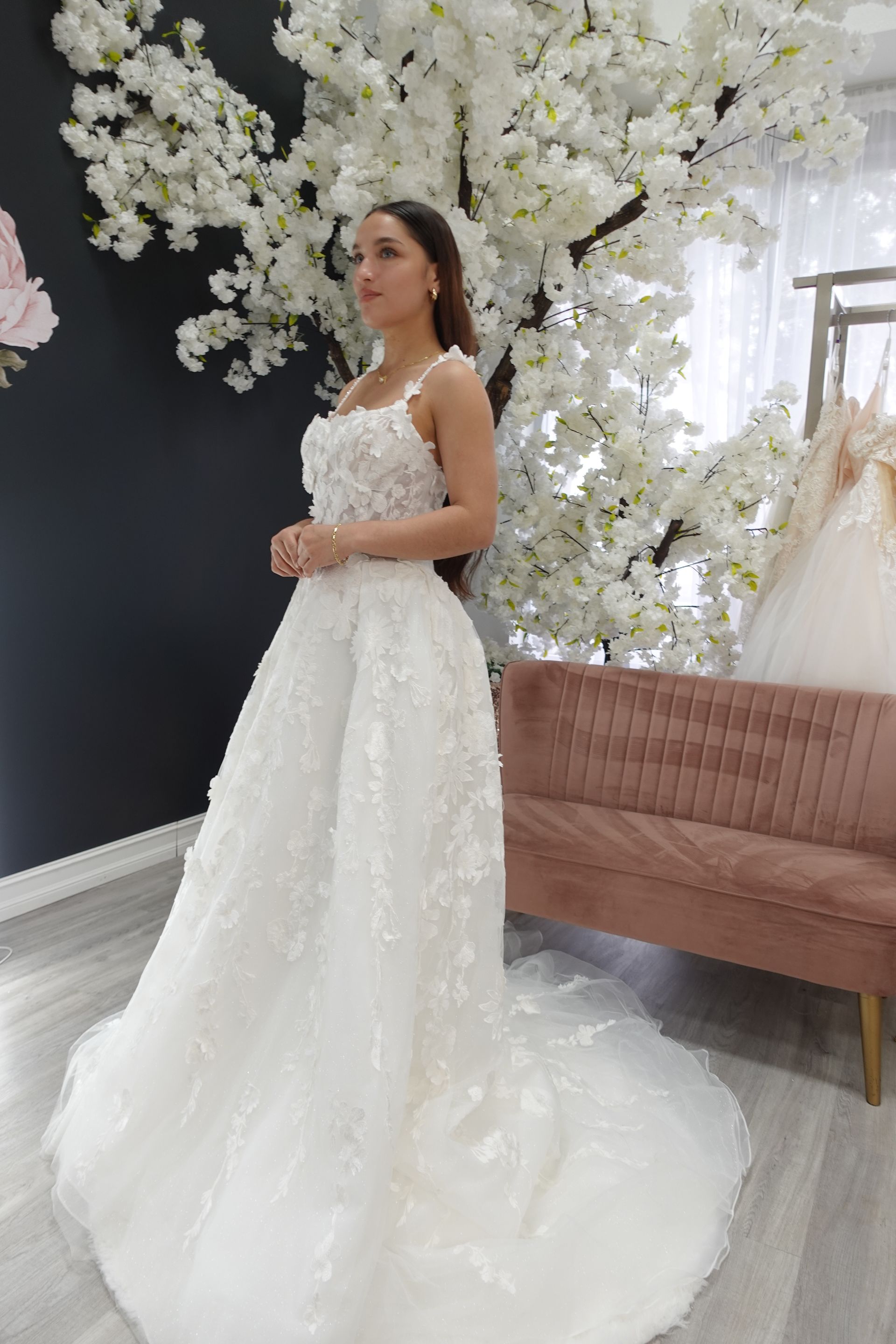 Woman in a white wedding dress stands near a floral backdrop and pink couch.