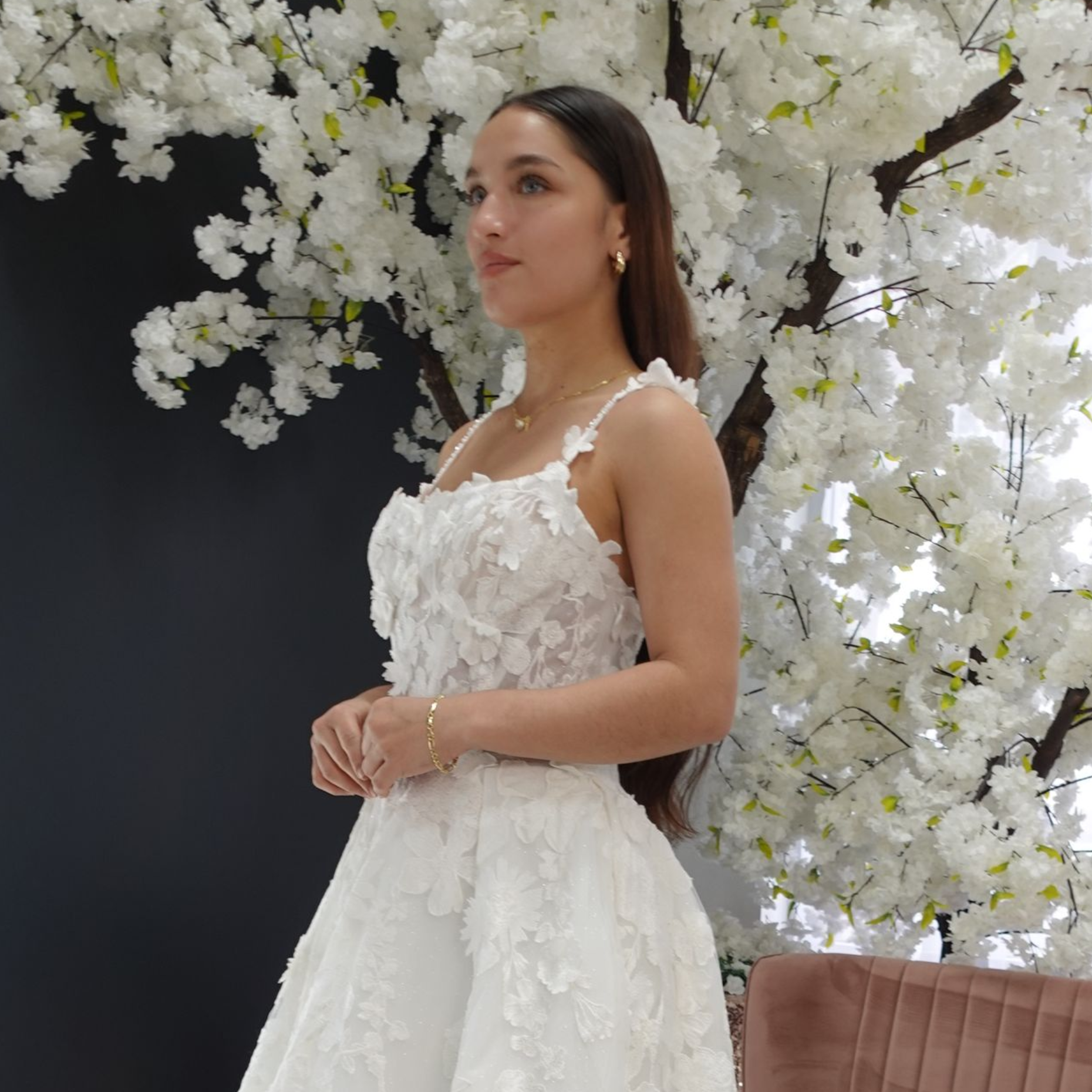 Woman in a white lace dress poses near white floral backdrop.