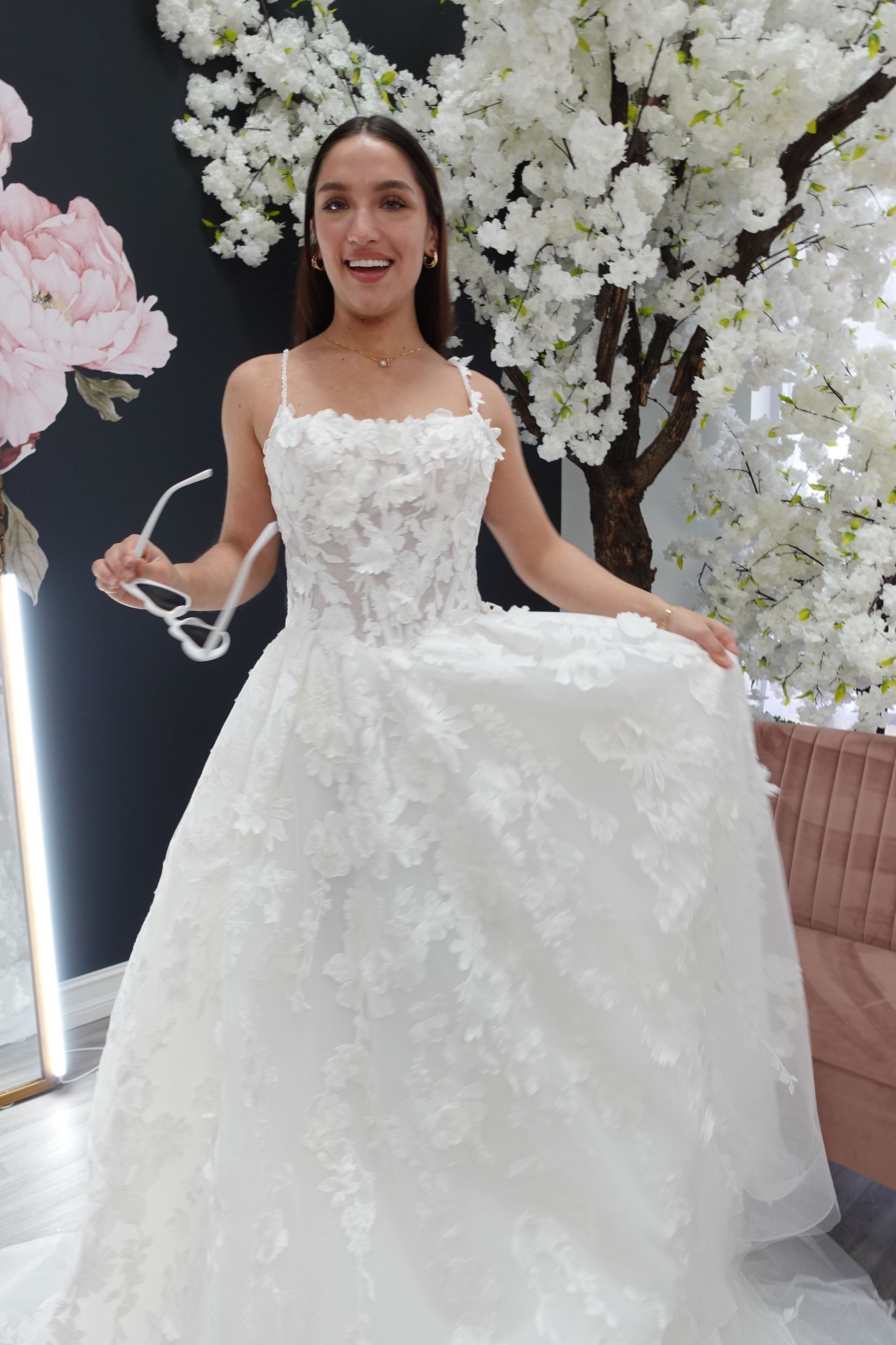 Woman in a white wedding dress with floral details, holding glasses, smiling in a bridal shop.
