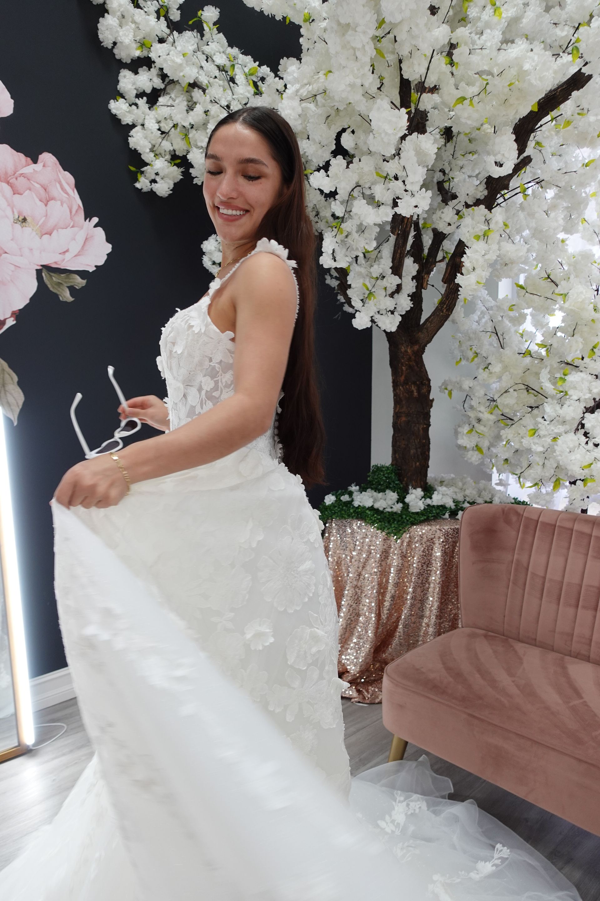 Woman in a white wedding dress stands in a bridal shop, near a floral backdrop and a pink sofa.