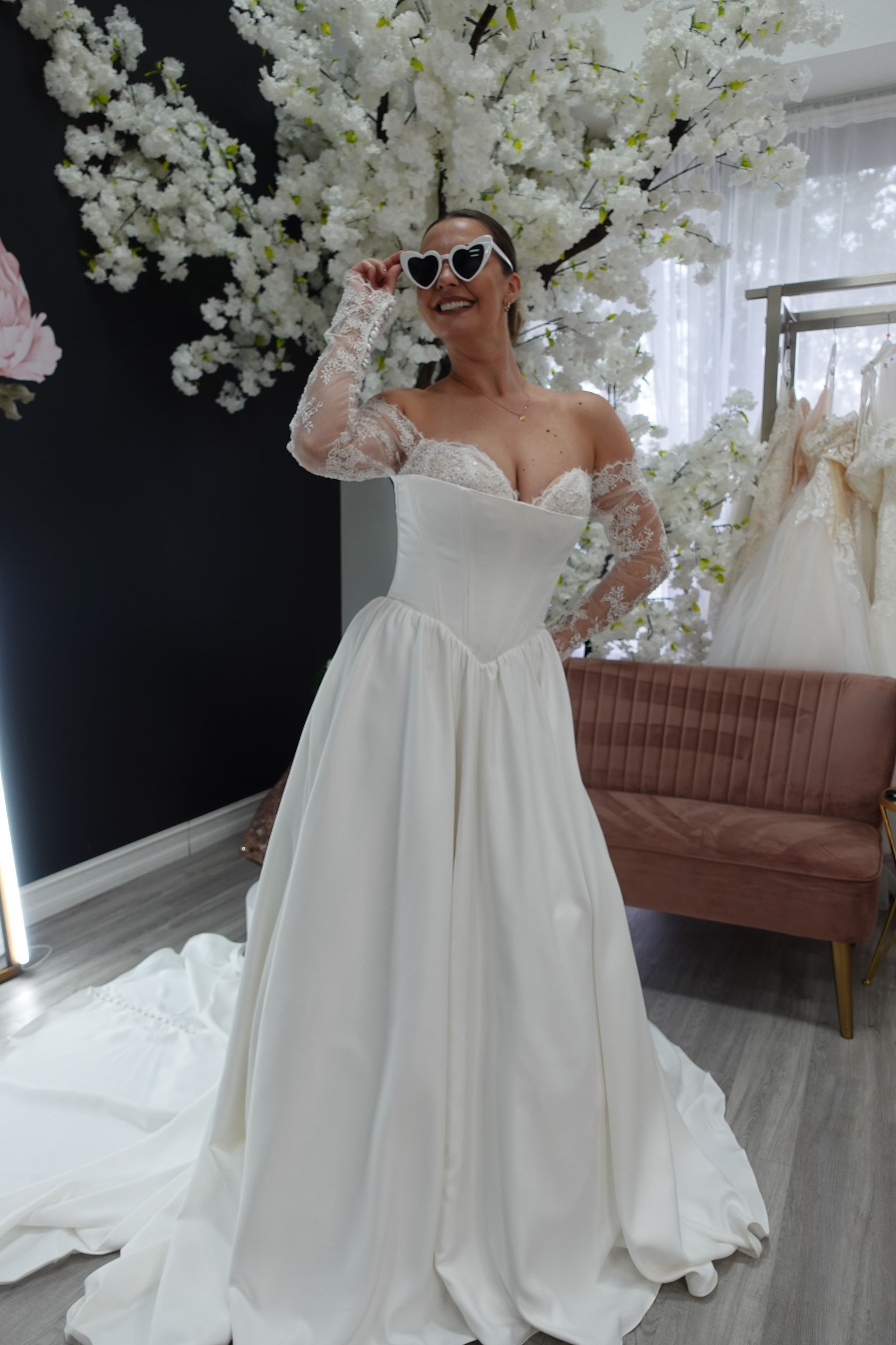 Woman in white off-the-shoulder wedding dress with long lace sleeves, smiles in a bridal shop.
