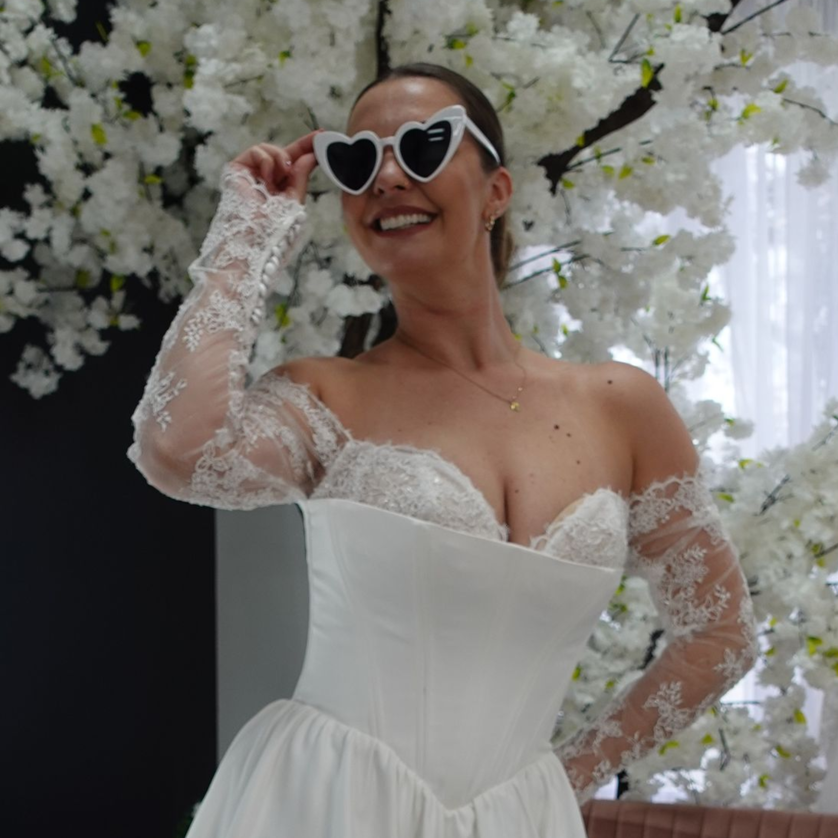 Woman in a white wedding dress with heart-shaped sunglasses, smiling. Posing in front of white flowers.
