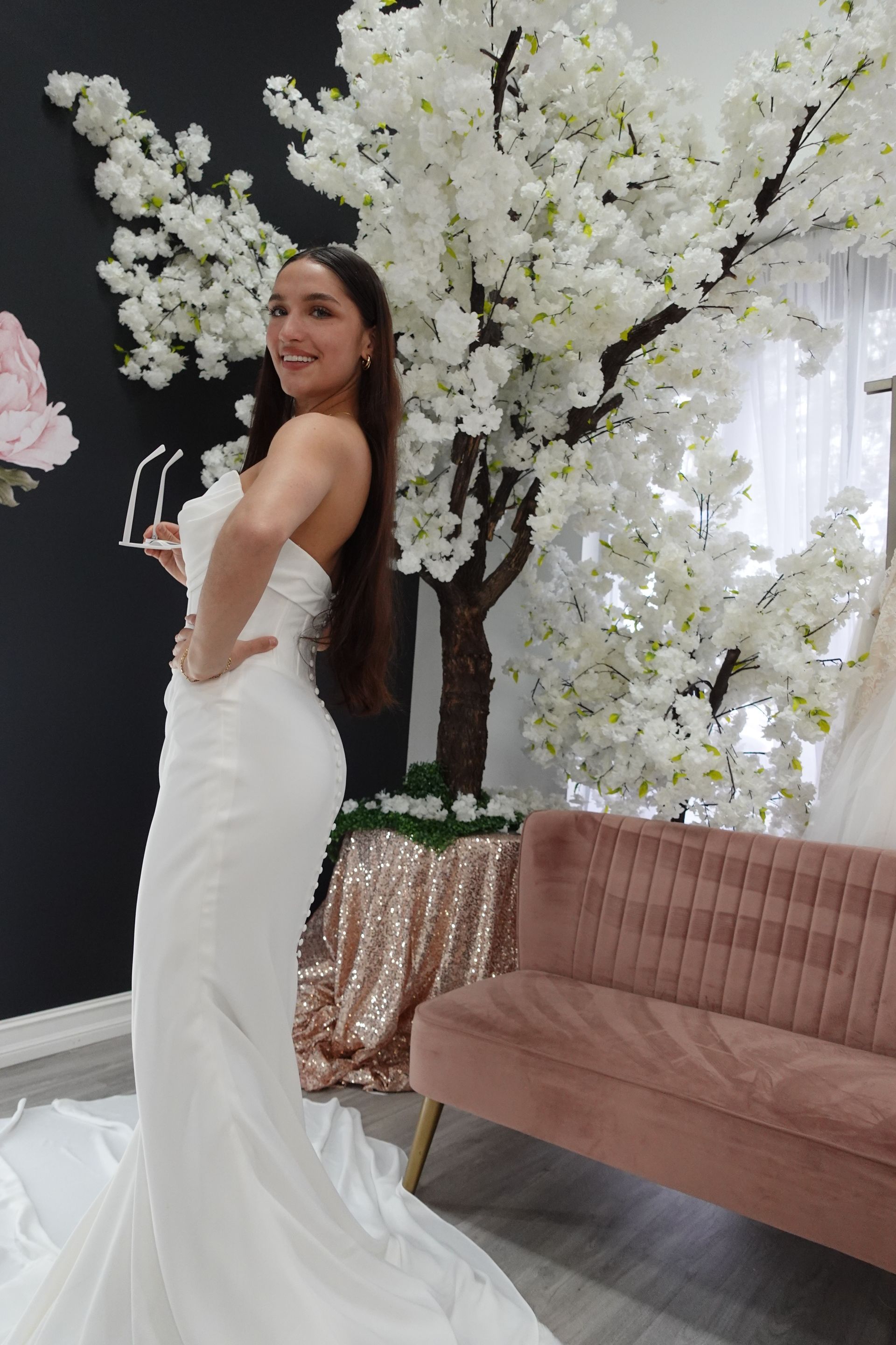 Woman in a white strapless gown poses, holding sunglasses, near a faux tree and pink couch.