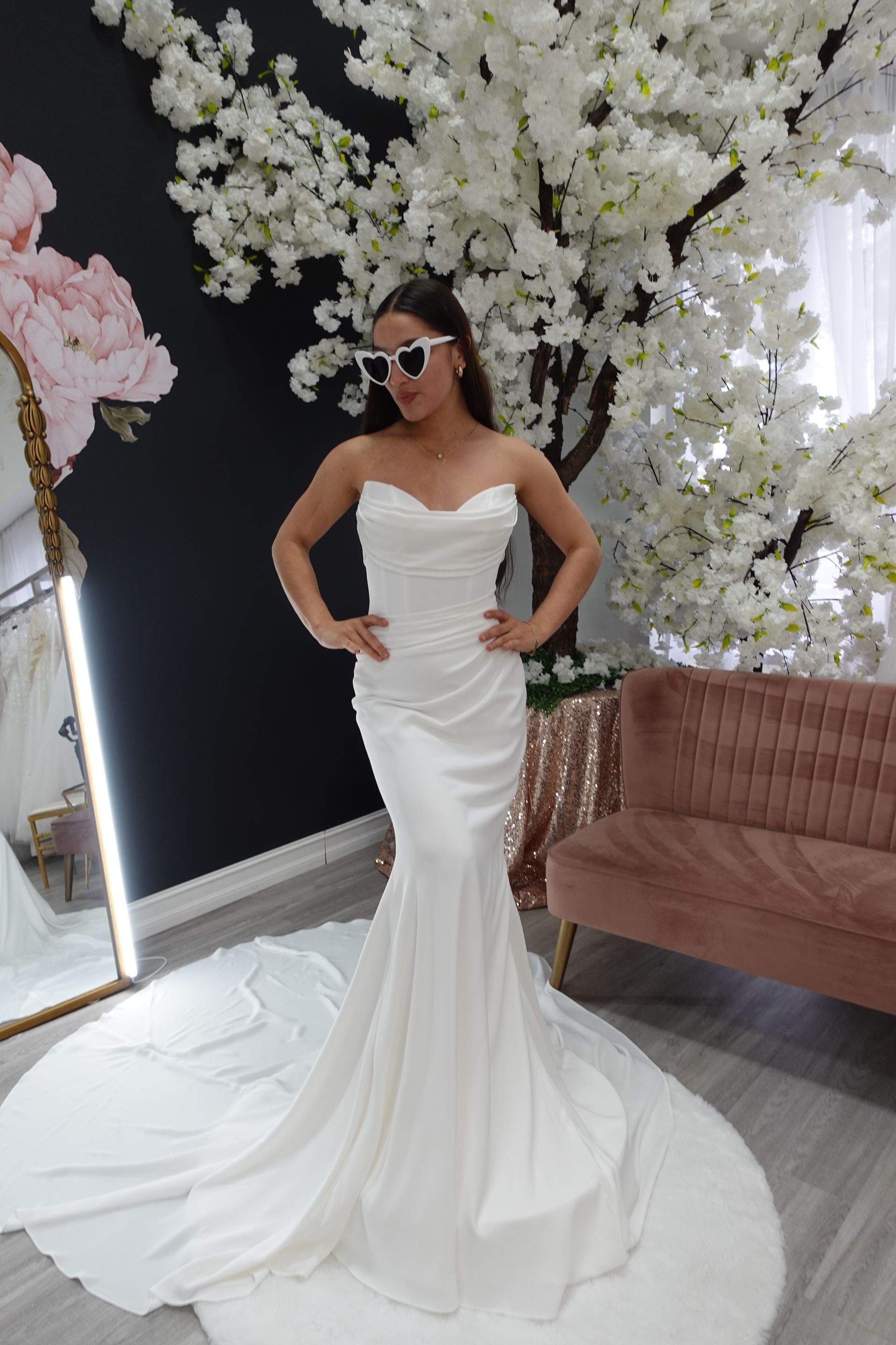 Woman in a white strapless mermaid wedding dress, posing with sunglasses in a shop, white floral backdrop.