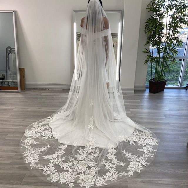 Woman in a white wedding dress with a long, lace-trimmed veil, standing in a bridal shop.
