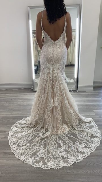 Woman in a lace wedding dress with a long train, standing in front of a mirror in a bridal shop.
