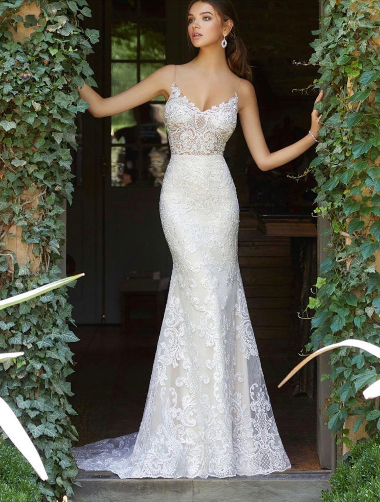 Woman in a white lace mermaid gown stands in an arched doorway, arms outstretched.