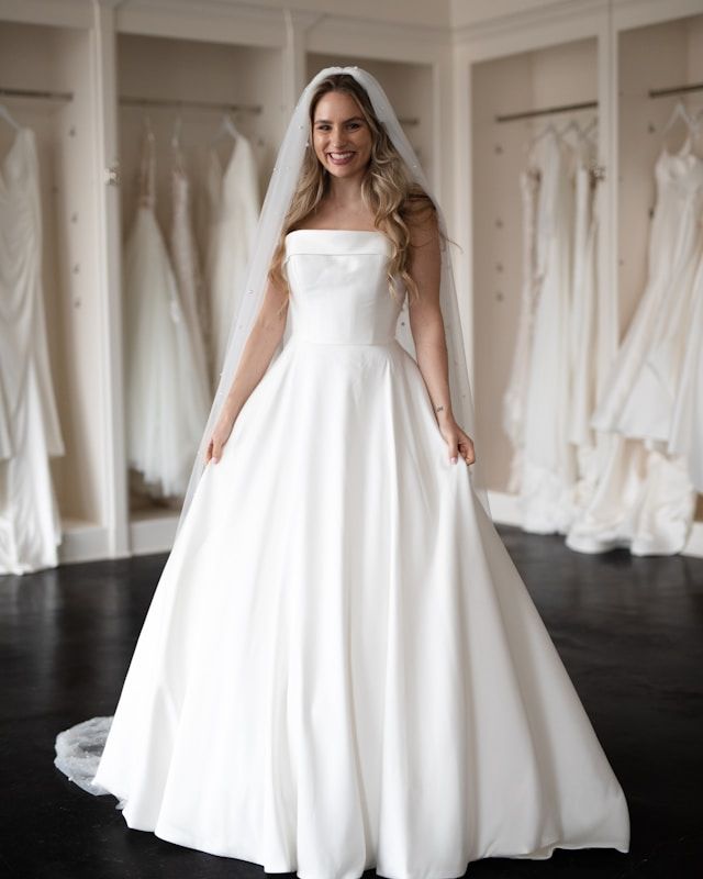 Woman in white wedding dress, holding the skirt, smiles in a bridal shop.