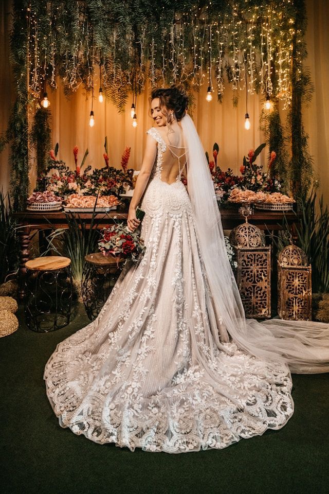 Bride in lace gown stands near a decorated table, back open, veil trailing. String lights and greenery in background.