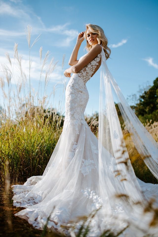 Woman in a white lace wedding dress poses in a field under a blue sky, holding her arm up.