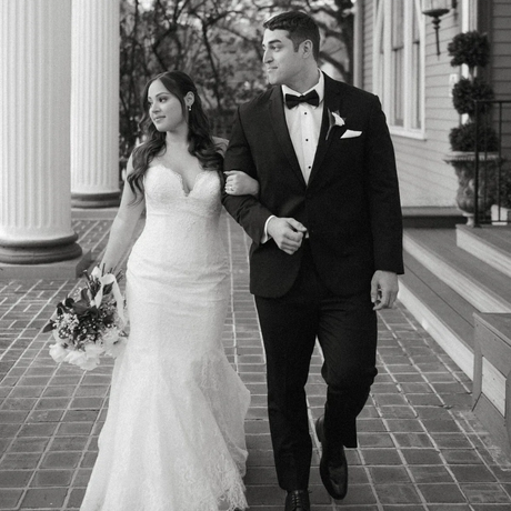 Bride and groom walking arm-in-arm, smiling. Outdoor, black and white. Bride in white gown, holding bouquet. Groom in tuxedo.