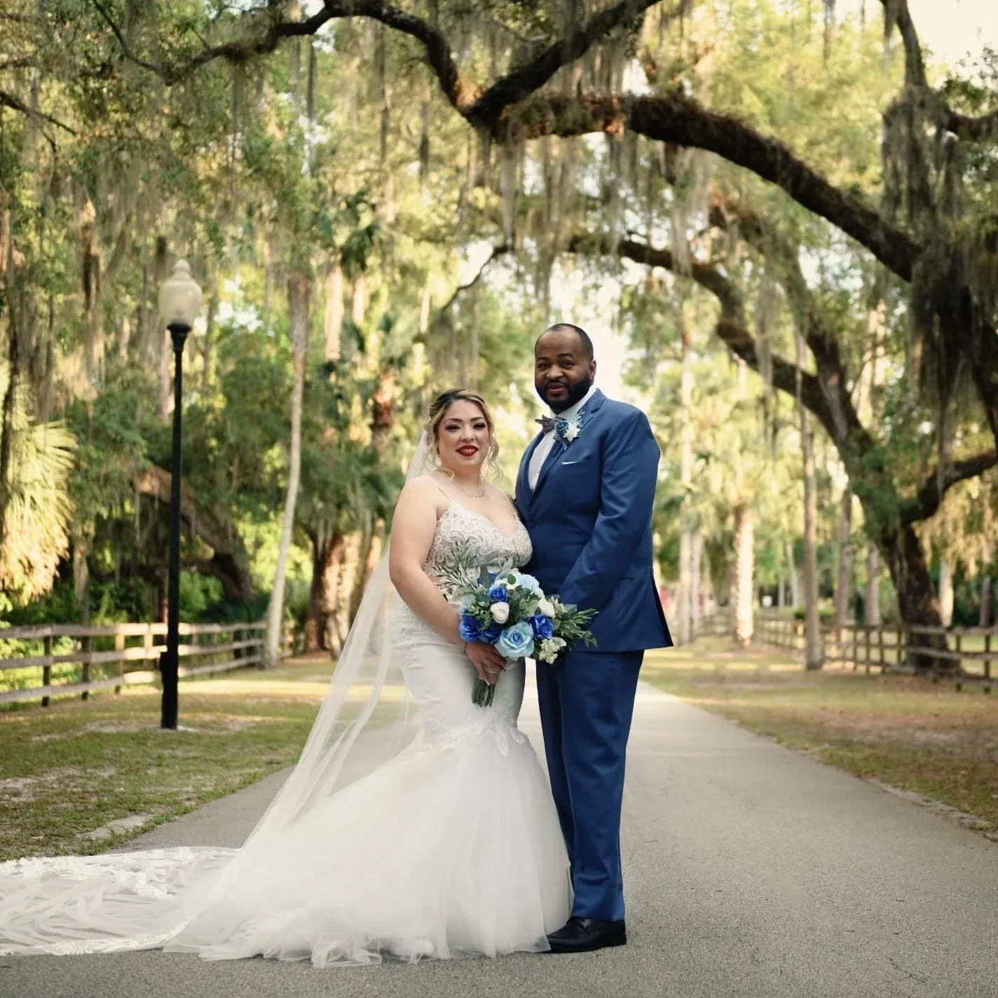 Bride and groom pose on path. Bride in white dress, groom in blue suit, with trees and fencing.