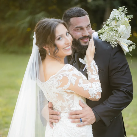 Bride and groom embrace outdoors; bride smiles, wearing lace dress and veil; groom with beard, wearing a suit.
