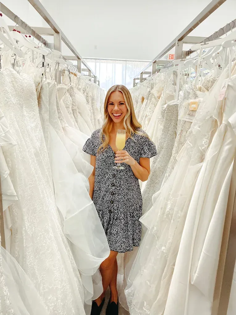 Woman in a black and white dress, holding a mimosa, smiles in a bridal shop, surrounded by wedding dresses.