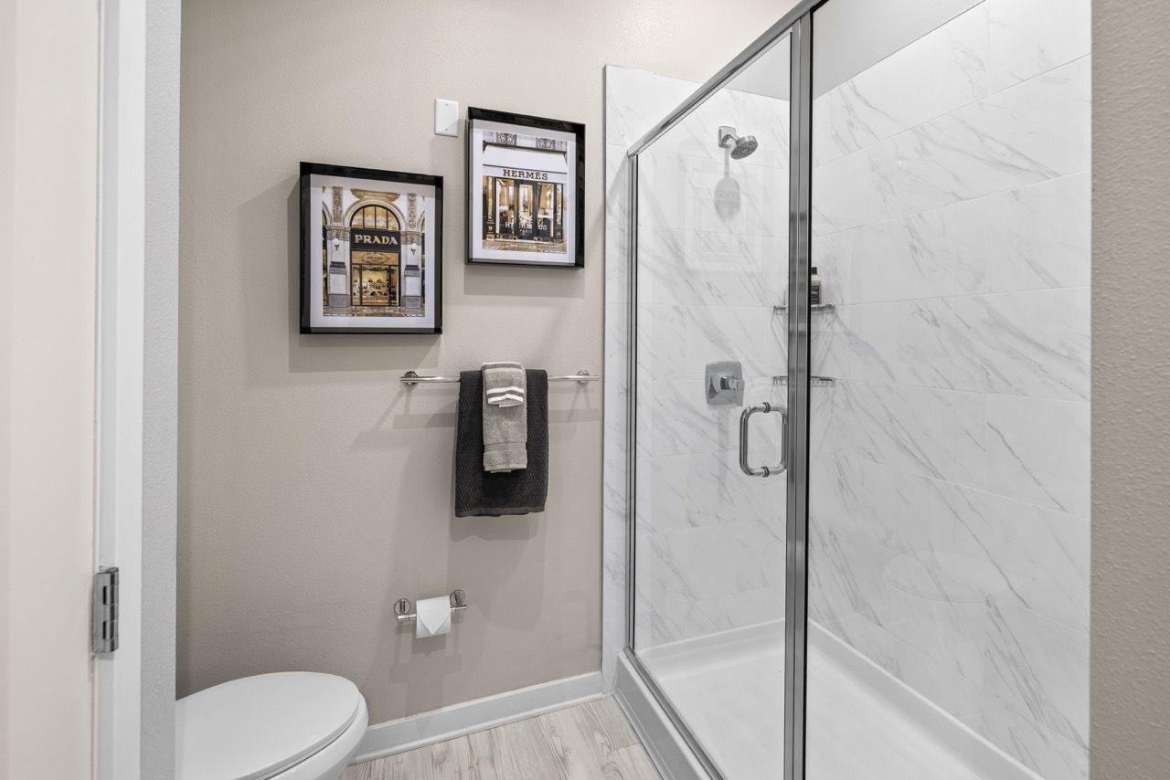 Bathroom interior with a glass-enclosed shower, toilet, towel bar, and framed art on beige wall.