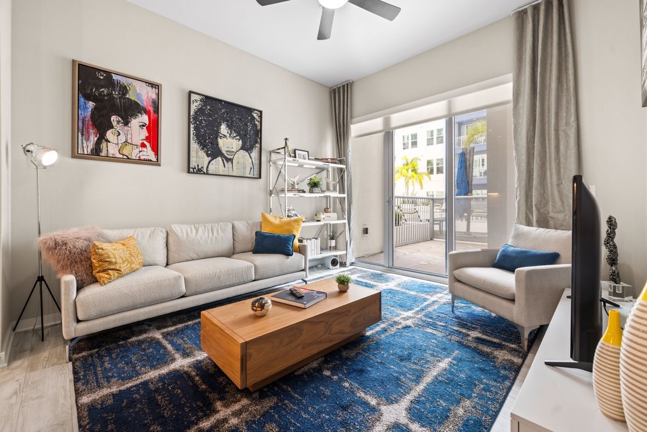Living room in an apartment with a beige sofa, armchair, coffee table, shelf unit, and sliding glass door to a balcony.