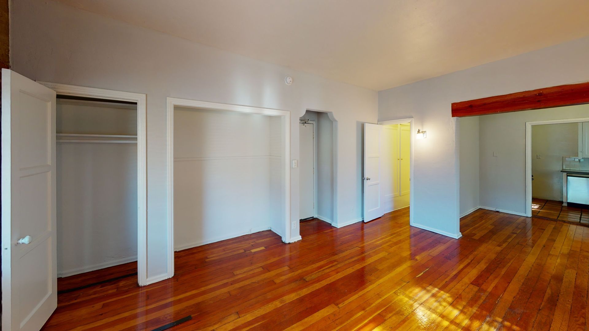 Empty room with hardwood floors, white walls, and open closets; a doorway leads to a kitchen.