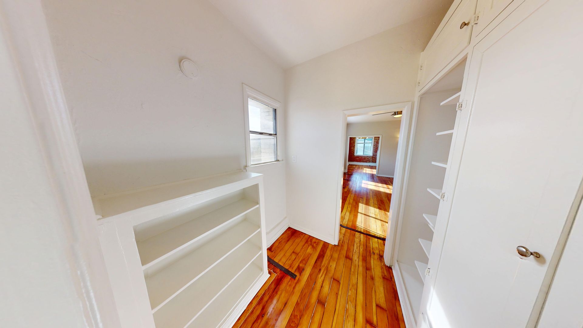 Narrow hallway with hardwood floors, built-in shelving and cabinets, leading to a room with sunlight.