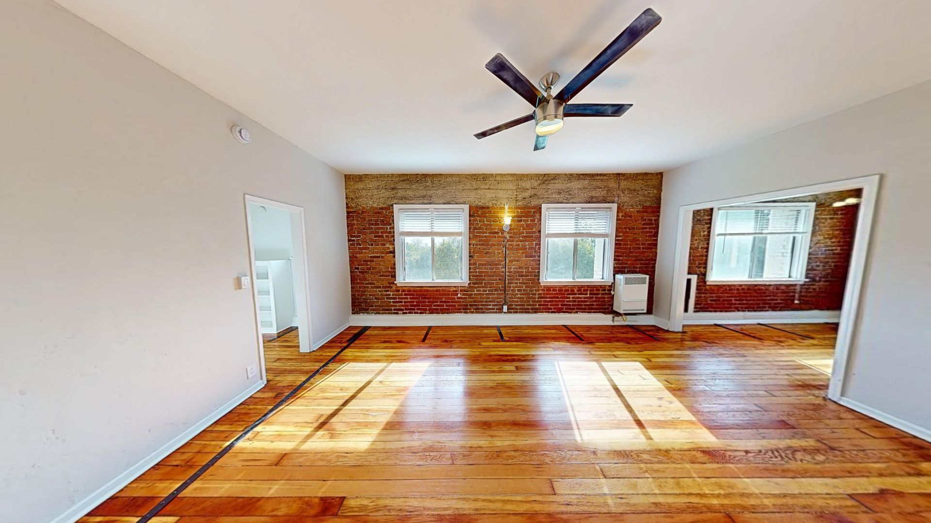 Empty living space with wood floor, brick wall, windows, and ceiling fan.