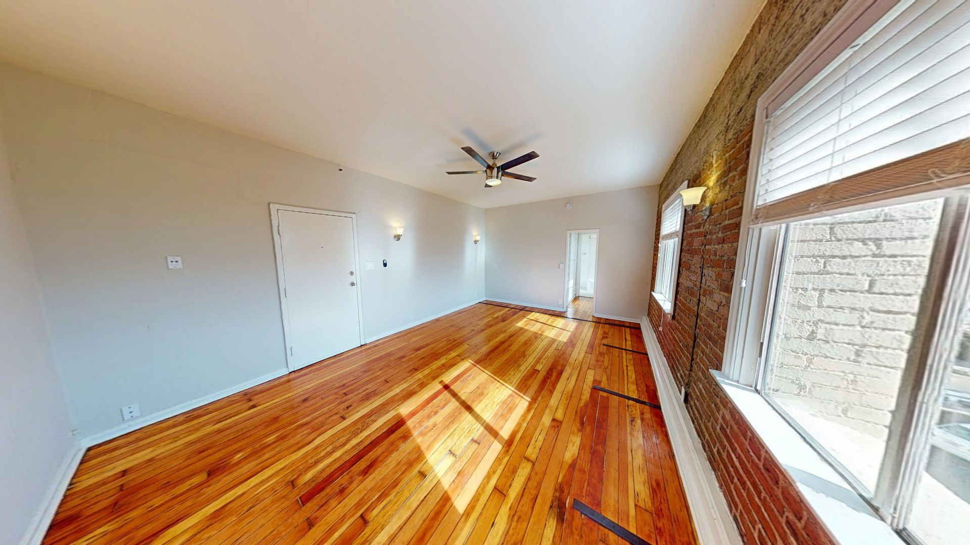 Empty room with wood floor, exposed brick wall, windows with blinds, and a ceiling fan.