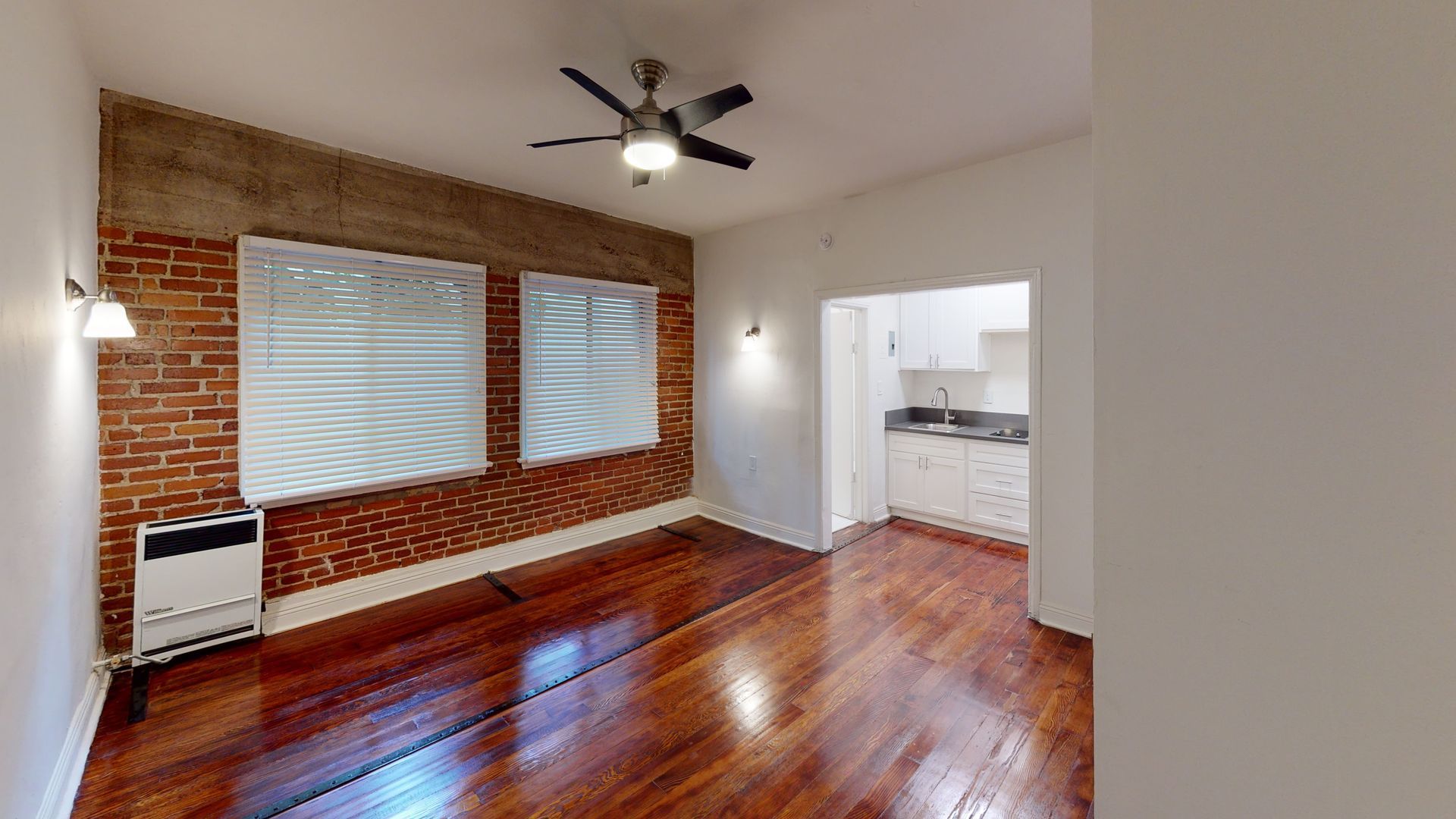 Interior of a room with wood floors, brick wall, and small kitchen visible.