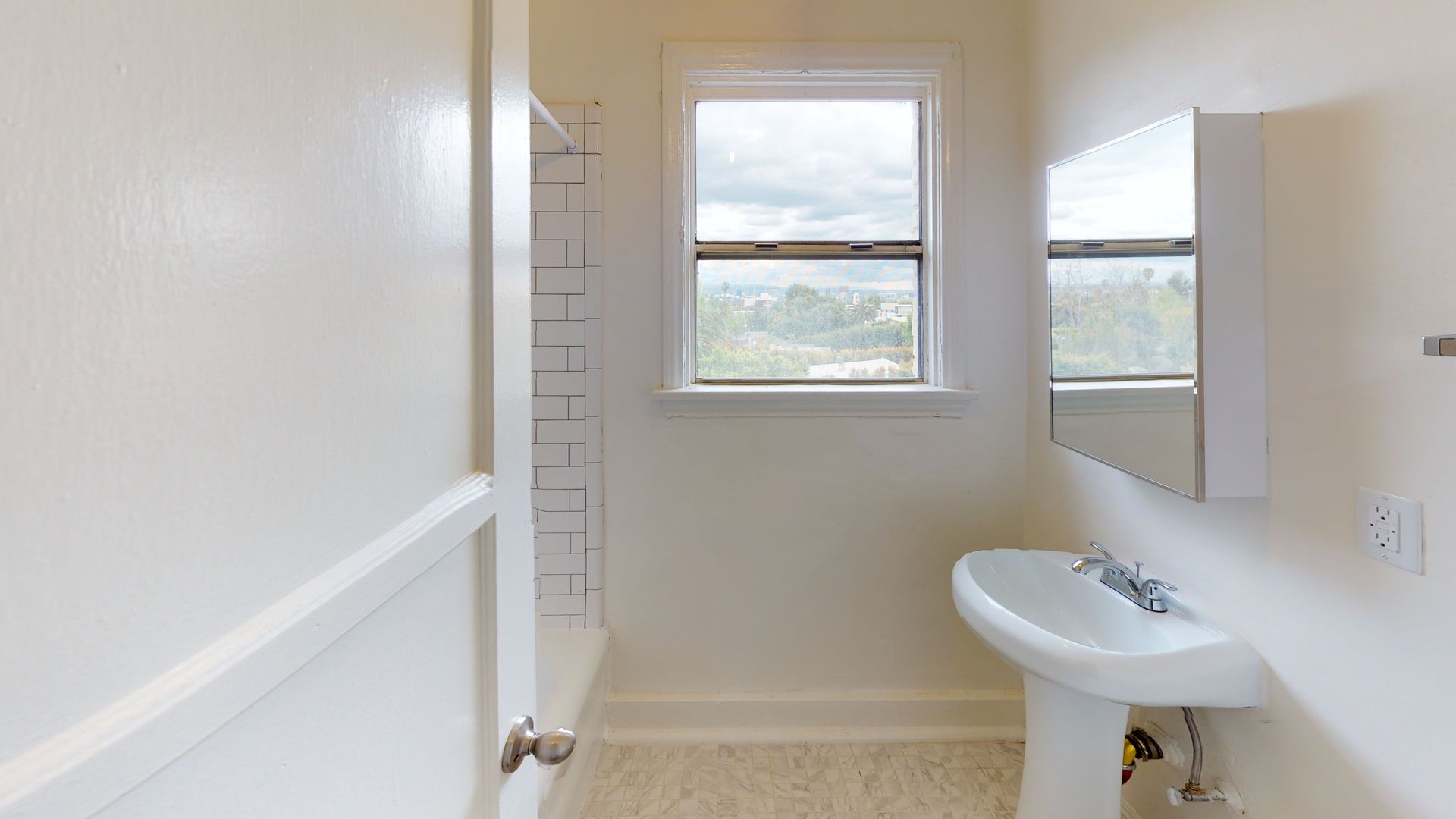 Bathroom with a window, sink, mirror, and shower. The walls and fixtures are white.