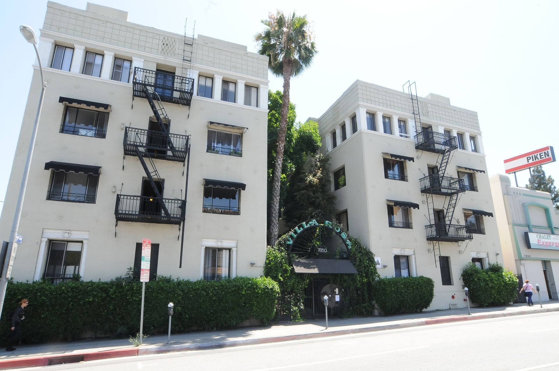 Two light-colored apartment buildings with black fire escapes, entrance covered in greenery, palm tree in between.