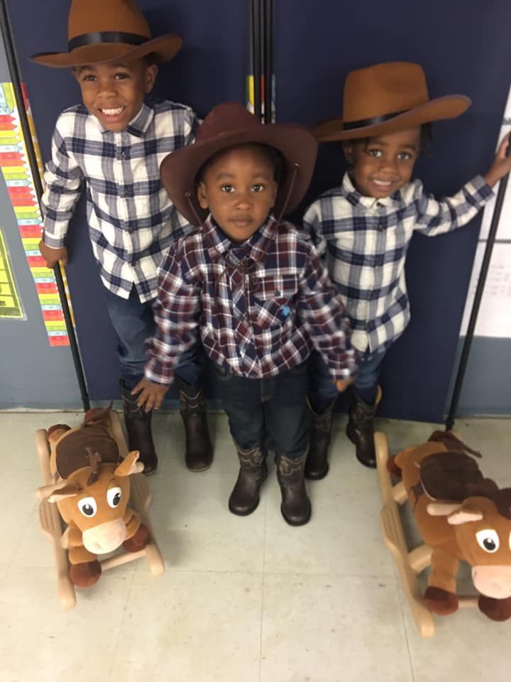 Three young boys dressed as cowboys are standing next to rocking horses.